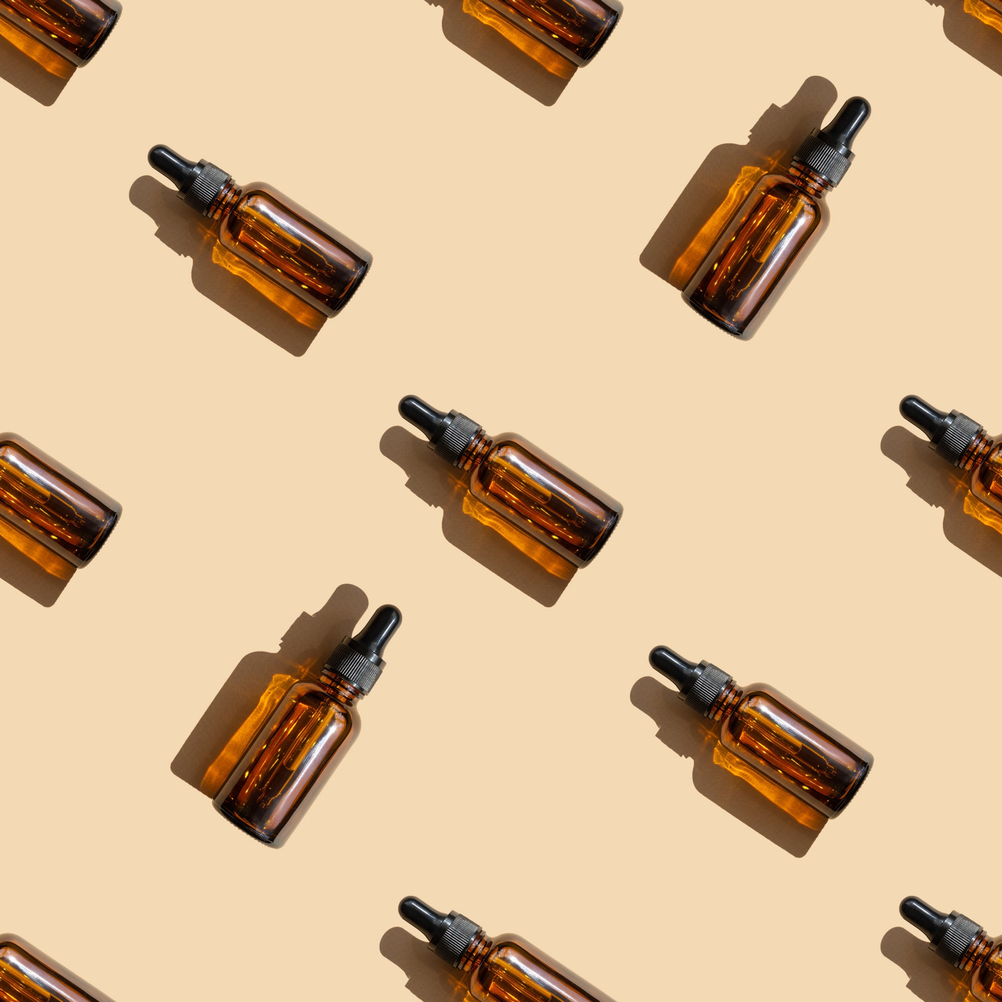 Seamless pattern of mock-ups of amber-colored glass bottles with dropper lids on beige background. Unmarked package from facial serum with a hard shadow cast, located randomly.