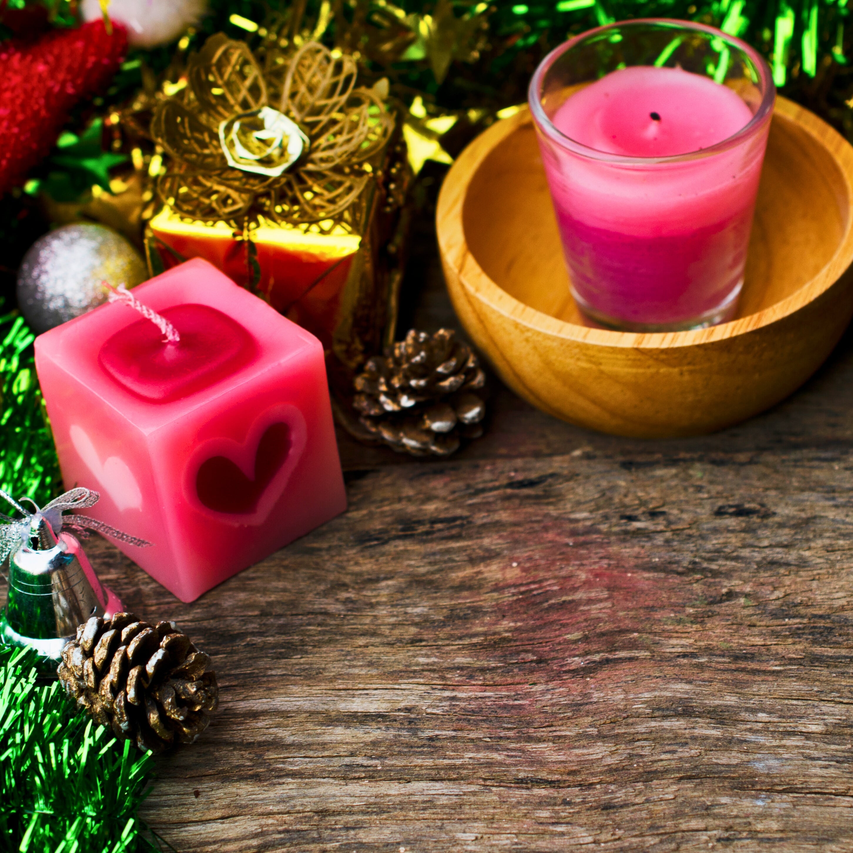 Two pink candles, one square with a heart, sit among pinecones and festive greenery on a weathered wooden surface.