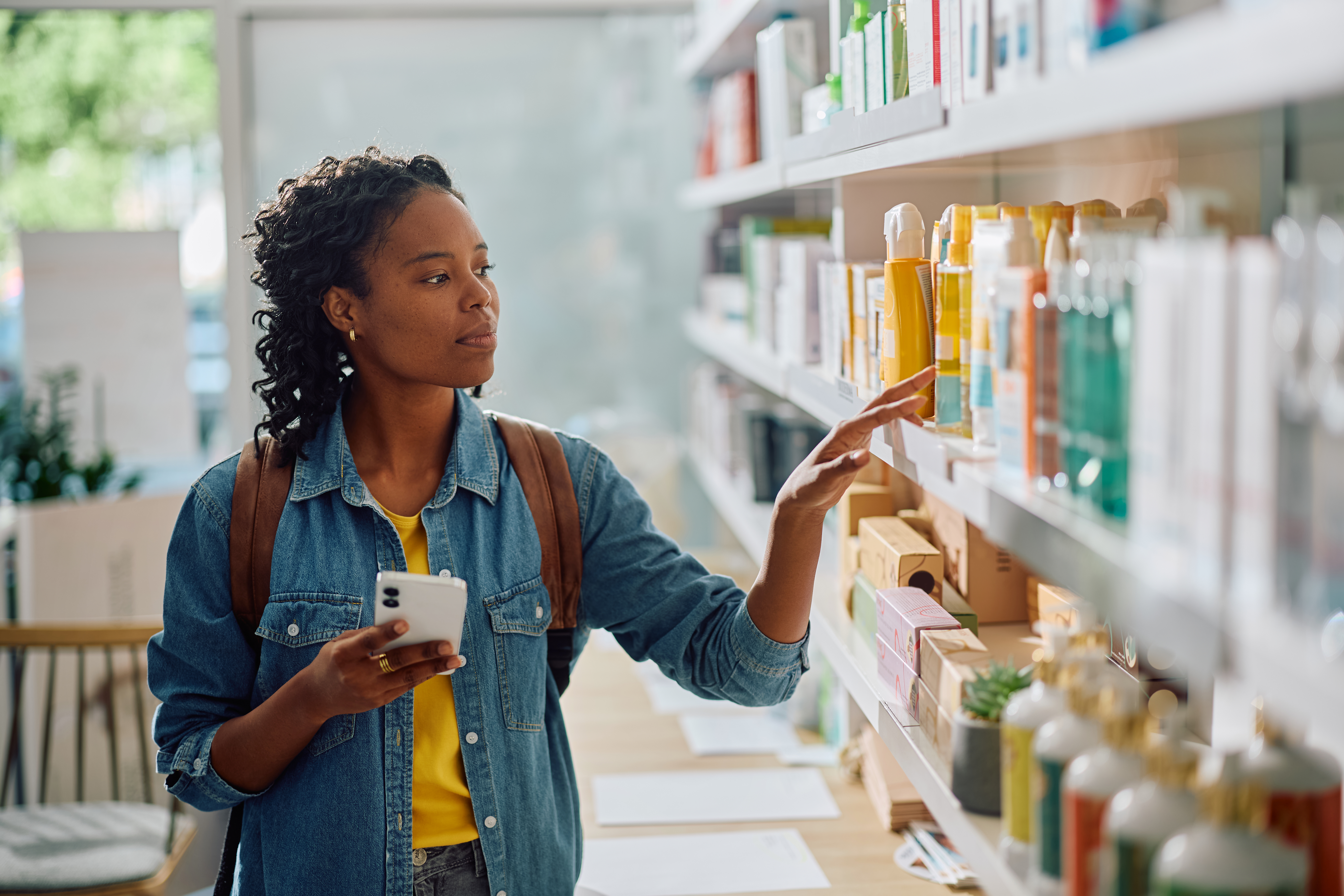 African American woman using mobile phone while looking for body care products at drugstore.